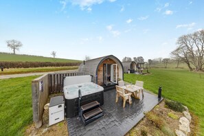 Un espace extérieur accueillant avec un jacuzzi, une terrasse en bois, une table avec des chaises et un petit bâtiment en bois dont la porte est ouverte.