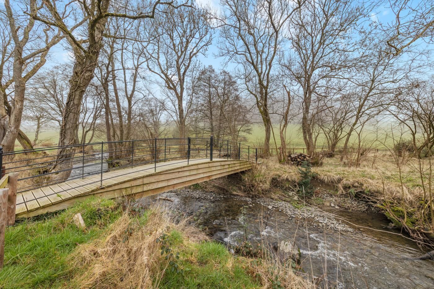 A wooden bridge over a stream with a metal railing, surrounded by trees and grassy areas.