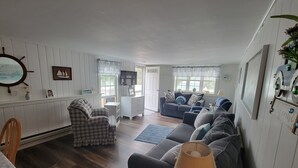 A cozy living room with a checkered armchair, a dark grey sofa, and a lamp. There is a white dresser and a window with curtains. - 103 North Shore Blvd Unit 1 (Sandwich)