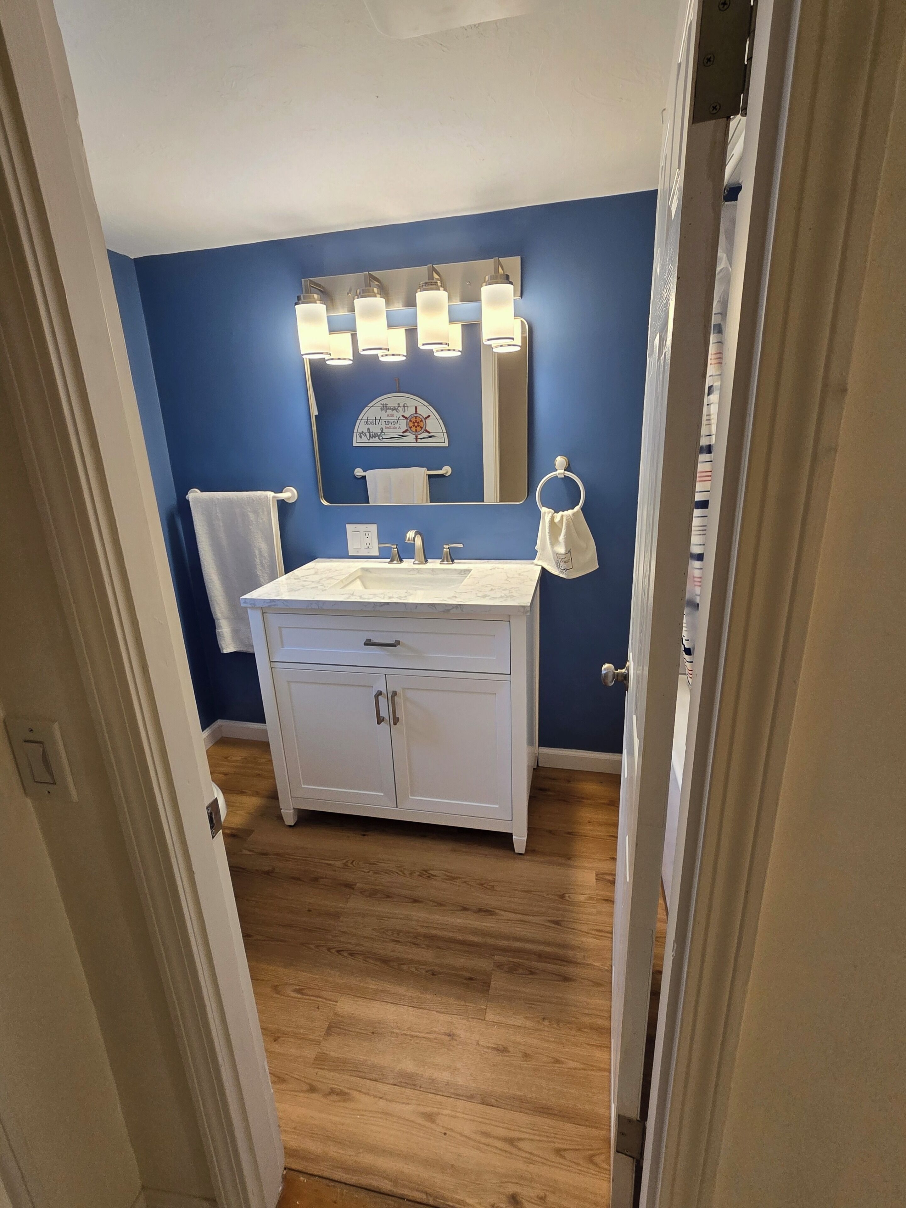 Een badkamer met een wit wastafelmeubel, een spiegel met verlichting, een handdoekrek en een houten vloer.