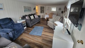 A cozy living room with a grey sofa, a blue armchair, and a matching ottoman. A dining table with chairs is in the background, and a TV is mounted on the wall. - 103 North Shore Blvd Unit 1 (Sandwich)