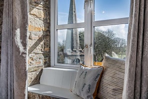 A stone wall, a window with a view of a church spire, and a bench with patterned cushions.