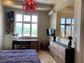 A bedroom with a bed, a desk, a chair, a dresser, a TV, and a large window with a view of greenery and buildings. - Riverside Gem in the center of Budapest is two-bedroom, cozy and comfortable (Budapest)