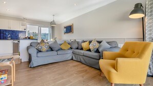 A modern living room with a grey sectional sofa, a yellow armchair, and a kitchen in the background. - 3 Bed in St. Merryn (oc-l29062) (Saint Merryn)