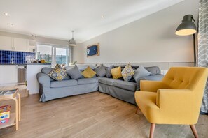 Een moderne woonkamer met een grijze modulaire bank, een gele fauteuil en een keuken op de achtergrond.