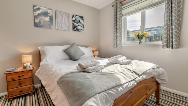 A neatly made bed with white linens and grey accents, a bedside table with a lamp, and a window with a view of buildings and flowers. - 3 Bed in St. Merryn (oc-l29062) (Saint Merryn)