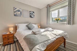A neatly made bed with white linens and grey accents, a bedside table with a lamp, and a window with a view of buildings and flowers. - 3 Bed in St. Merryn (oc-l29062) (Saint Merryn)