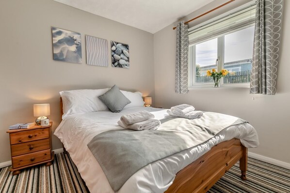 Een net opgemaakt bed met witte lakens en grijze accenten, een nachtkastje met een lamp, en een raam met uitzicht op gebouwen en bloemen.
