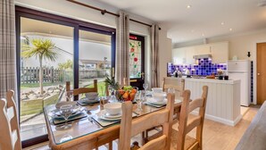 Uma sala de jantar com mesa posta para quatro, uma cozinha com armários brancos e azulejos azuis, e vista para um jardim por grandes janelas.