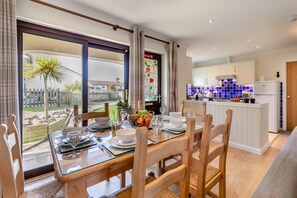 A dining area with a table set for four, a kitchen with white cabinets and blue tiles, and a view of a garden through large windows. - 2 Bed in St. Merryn (oc-l29063) (Treyarnon Bay)