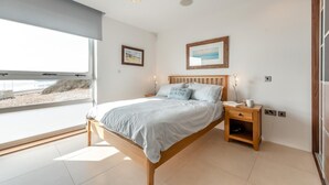 A bedroom with a bed, bedside tables, a window with a beach view, and a ceiling light. - 2 Bed in Tenby (oc-t29060) (Tenby)
