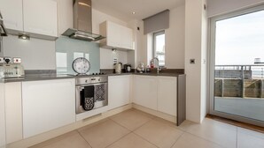 A modern kitchen with white cabinets, a built-in oven, a coffee maker, and a view of the sea through a large window. - 2 Bed in Tenby (oc-t29060) (Tenby)