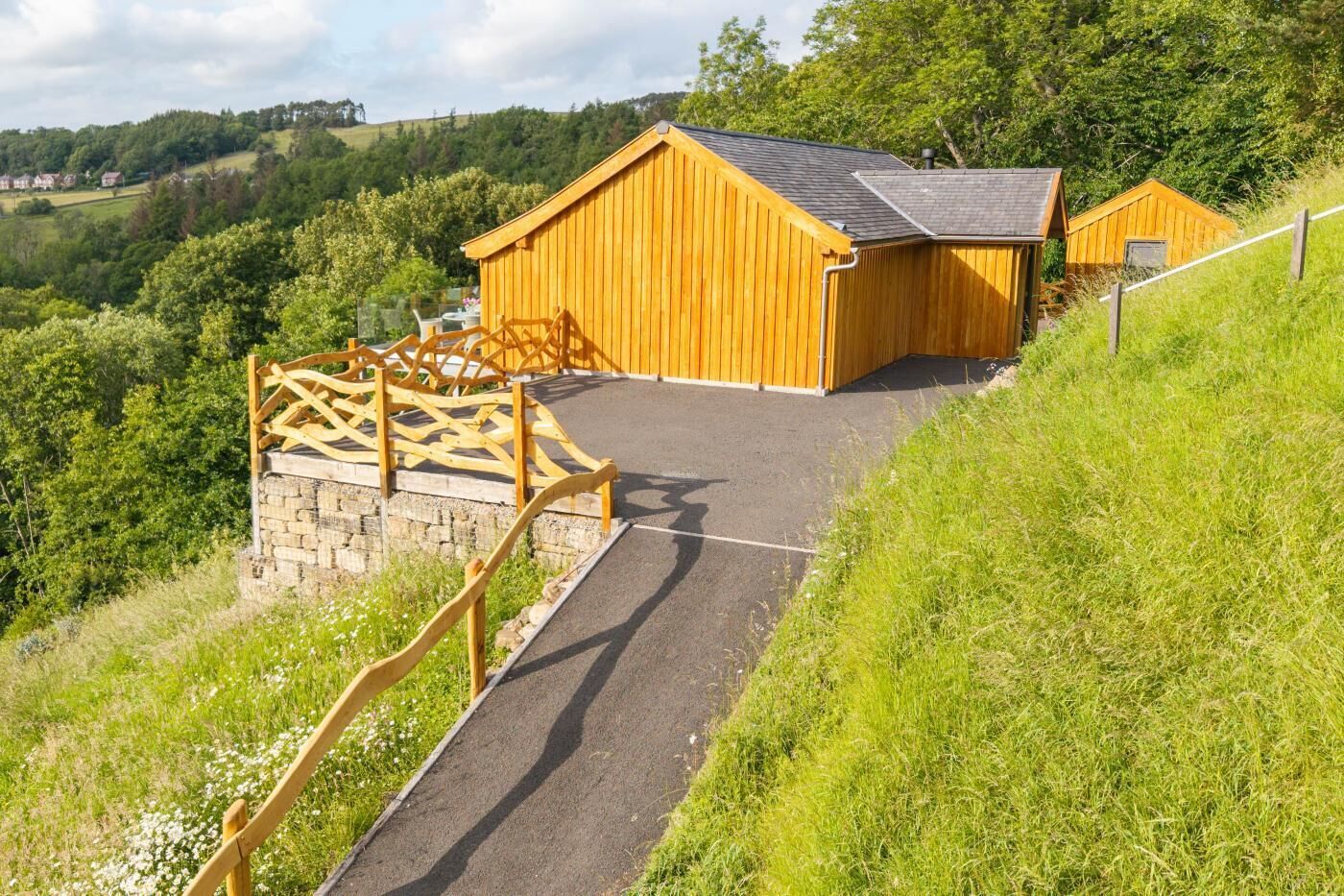 A wooden building with a sloped roof, a stone wall, and a wooden fence leading to a paved path.