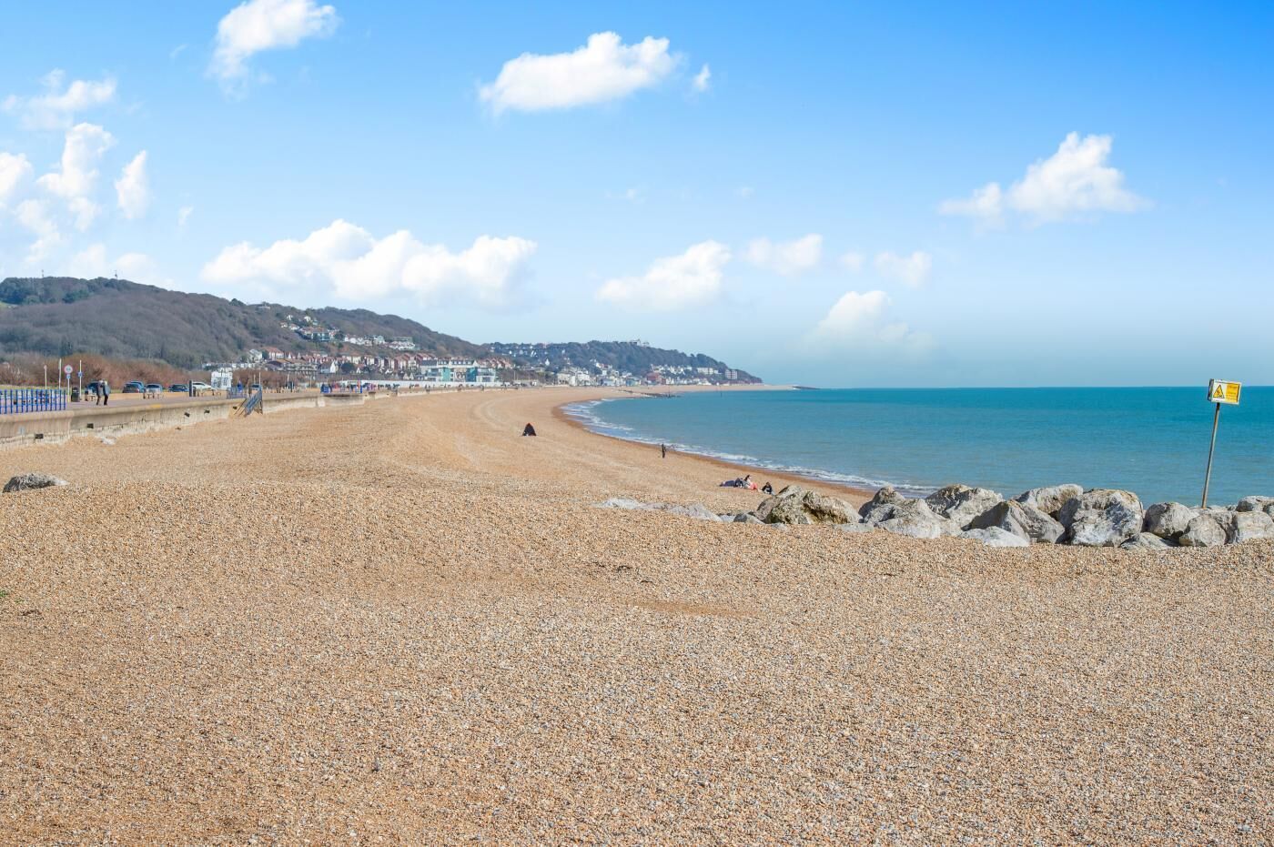Une plage de galets bordée d’une côte rocheuse, un ciel bleu dégagé et des bâtiments qui se dessinent à l’horizon.