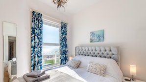 Quarto com cama grande, cortinas florais, vista para o mar, lustre e pintura na parede.
