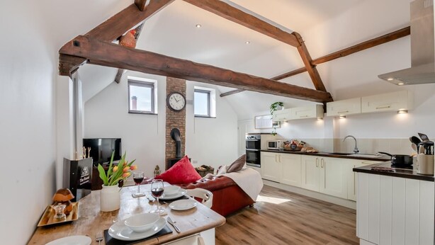 Una cocina moderna con armarios blancos, una encimera de madera y un horno empotrado. Hay una zona de comedor con una mesa de madera dispuesta para dos personas, y un banco con cojín rojo.