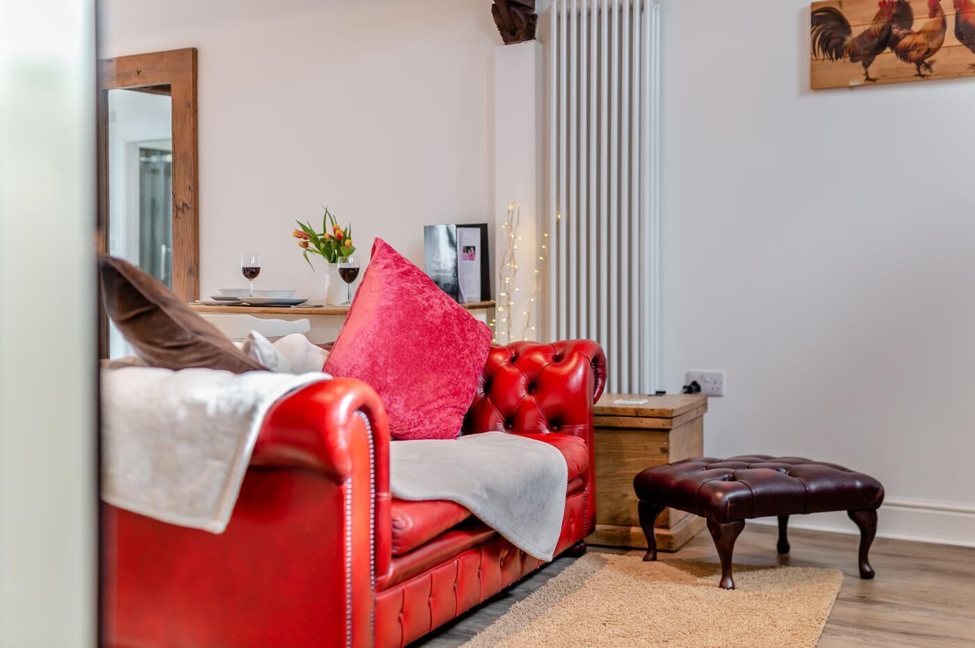 Ein rotes Ledersofa mit weißer Decke, einem roten Kissen und einem braunen Hocker in einem Wohnzimmer mit Holzboden, Heizkörper und einem gerahmten Bild von Hähnen an der Wand.
