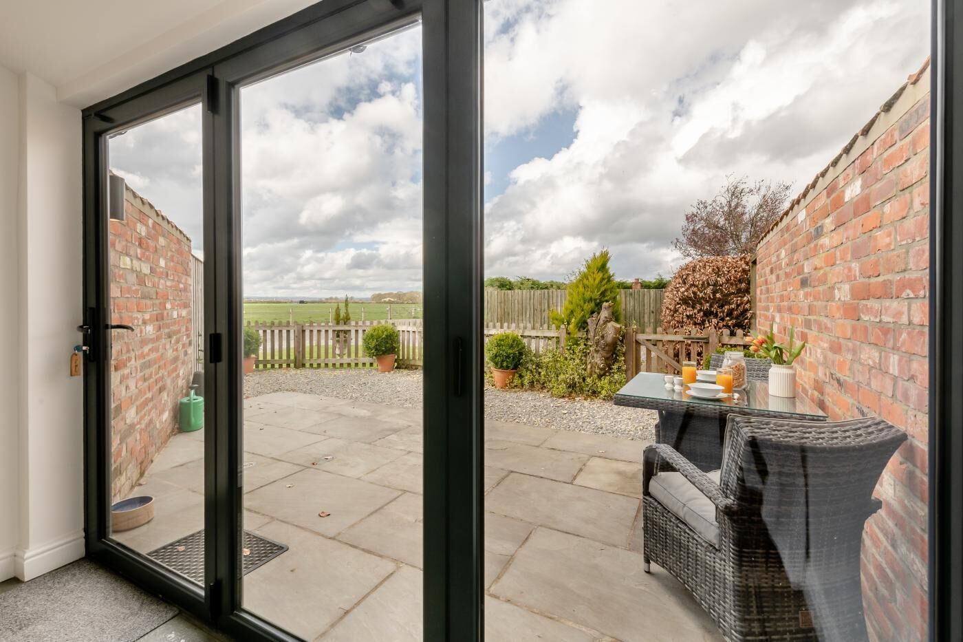 A patio with a glass door leading to a brick wall, a table with outdoor seating, and a view of a grassy field.