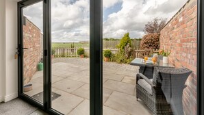 Un patio con puerta de vidrio que da a un muro de ladrillo, una mesa con asientos exteriores y vista a un campo cubierto de césped.
