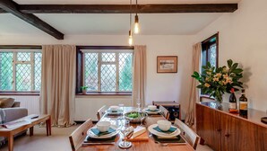 A dining area with a wooden table set for four, a window with curtains, a radiator, and a cabinet with flowers and bottles. - 2 Bed in Charing (oc-r29113) (Charing)
