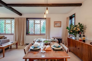 A dining area with a wooden table set for four, a window with curtains, a radiator, and a cabinet with flowers and bottles. - 2 Bed in Charing (oc-r29113) (Charing)