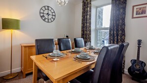 Een eetgedeelte met een houten tafel voor vier personen, zwarte lederen stoelen, een wandklok, een hangende kristallen lamp en een gitaar tegen de muur.