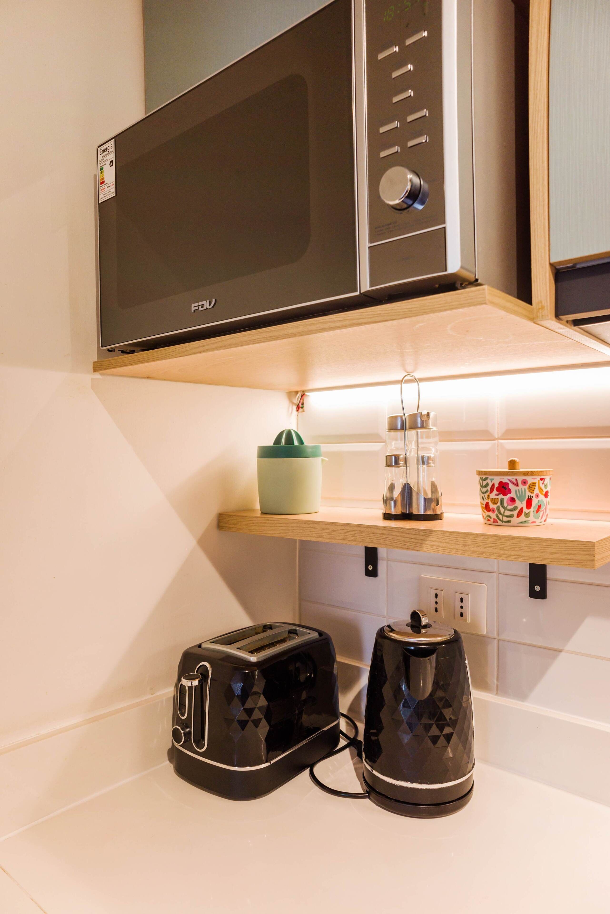 A kitchen with a microwave, toaster, kettle, and a shelf with a plant and a decorative pot.