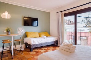 Ein Hotelzimmer mit Bett, Schreibtisch mit Ventilator, Fernseher und Blick auf Gebäude durch ein Fenster.
