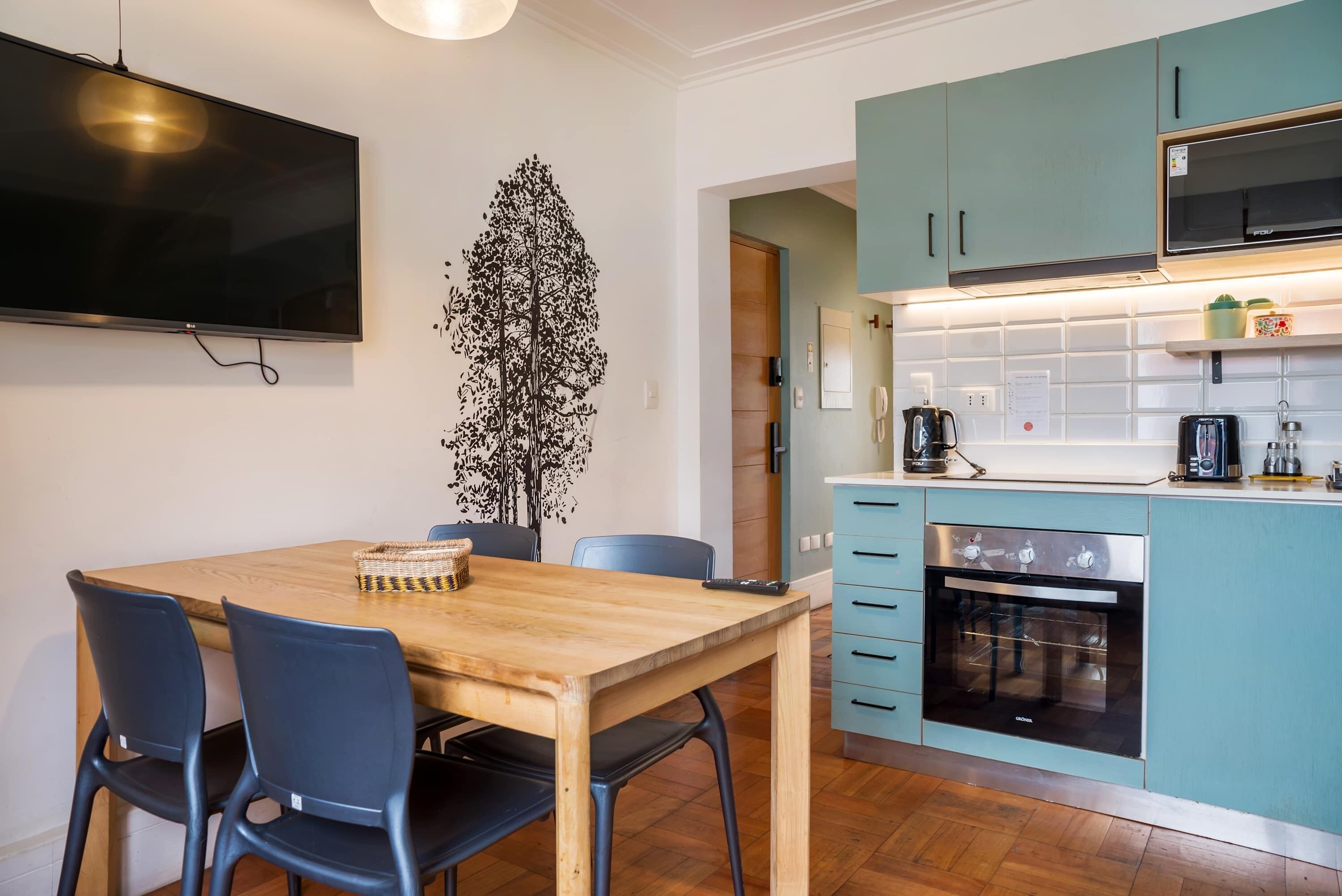 A modern kitchen with teal cabinets, a built-in oven, and a flat-screen TV mounted on the wall.