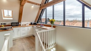 Eine Küche mit Holzboden, weißen Schränken, einer Holzausziehtheke und Blick zum Himmel durch große Fenster.