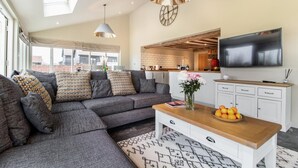 Ein modernes Wohnzimmer mit einer Eckcouch, einem Couchtisch mit Obst, einem Flachbildfernseher und einer Küche im Hintergrund.