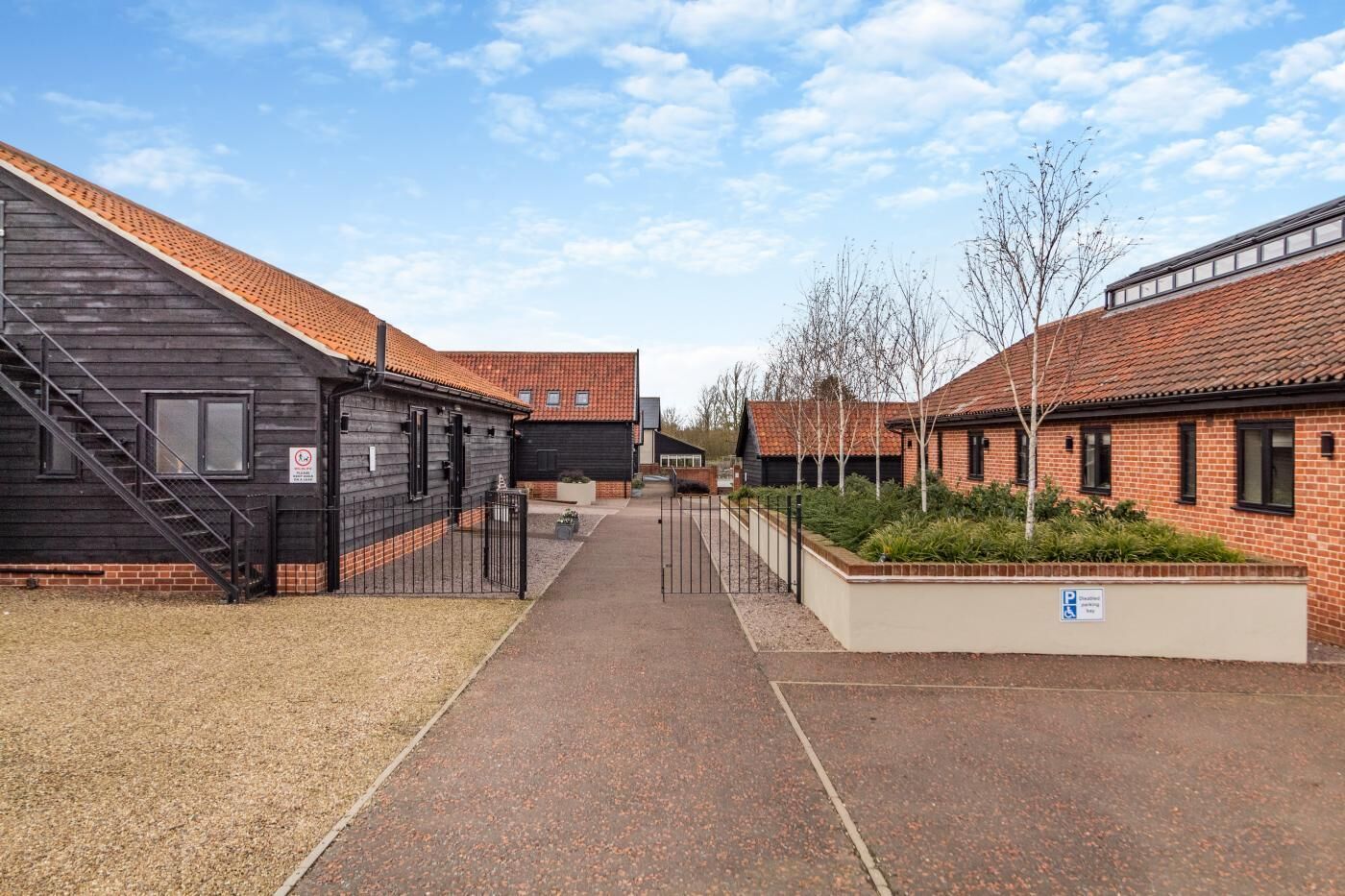 A courtyard with a paved walkway, a black building with a staircase, and a brick building with a garden area.