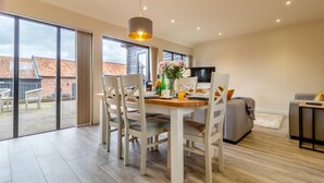 Un comedor moderno con una mesa dispuesta para dos, un sofá y un televisor en una habitación amplia con ventanas grandes y vistas a edificios.