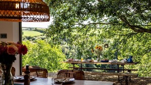 An outdoor dining area with a table set for two, surrounded by lush greenery and a scenic view. - 5 Bed in Little Petherick (oc-s29057) (Little Petherick)