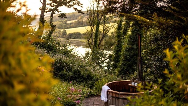 A wooden hot tub in a garden with a bench and a table, surrounded by lush greenery and a view of a lake. - 5 Bed in Little Petherick (oc-s29057) (Little Petherick)
