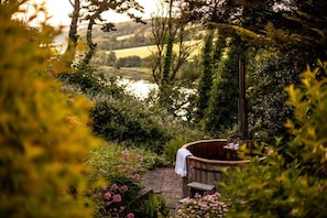 Uma banheira de hidromassagem de madeira num jardim, com um banco e uma mesa, rodeada por vegetação exuberante e vista para um lago.