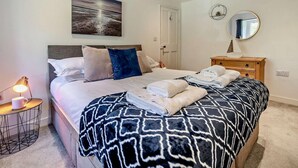 Una cama bien tendida con una manta estampada, una lámpara de noche, una mesita con una taza y un espejo fijado a la pared.