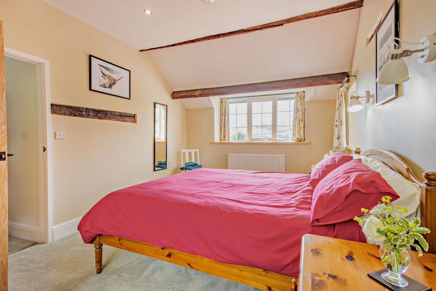 A bedroom with a wooden bed, a bedside table with a vase, a window with curtains, and a framed picture on the wall.