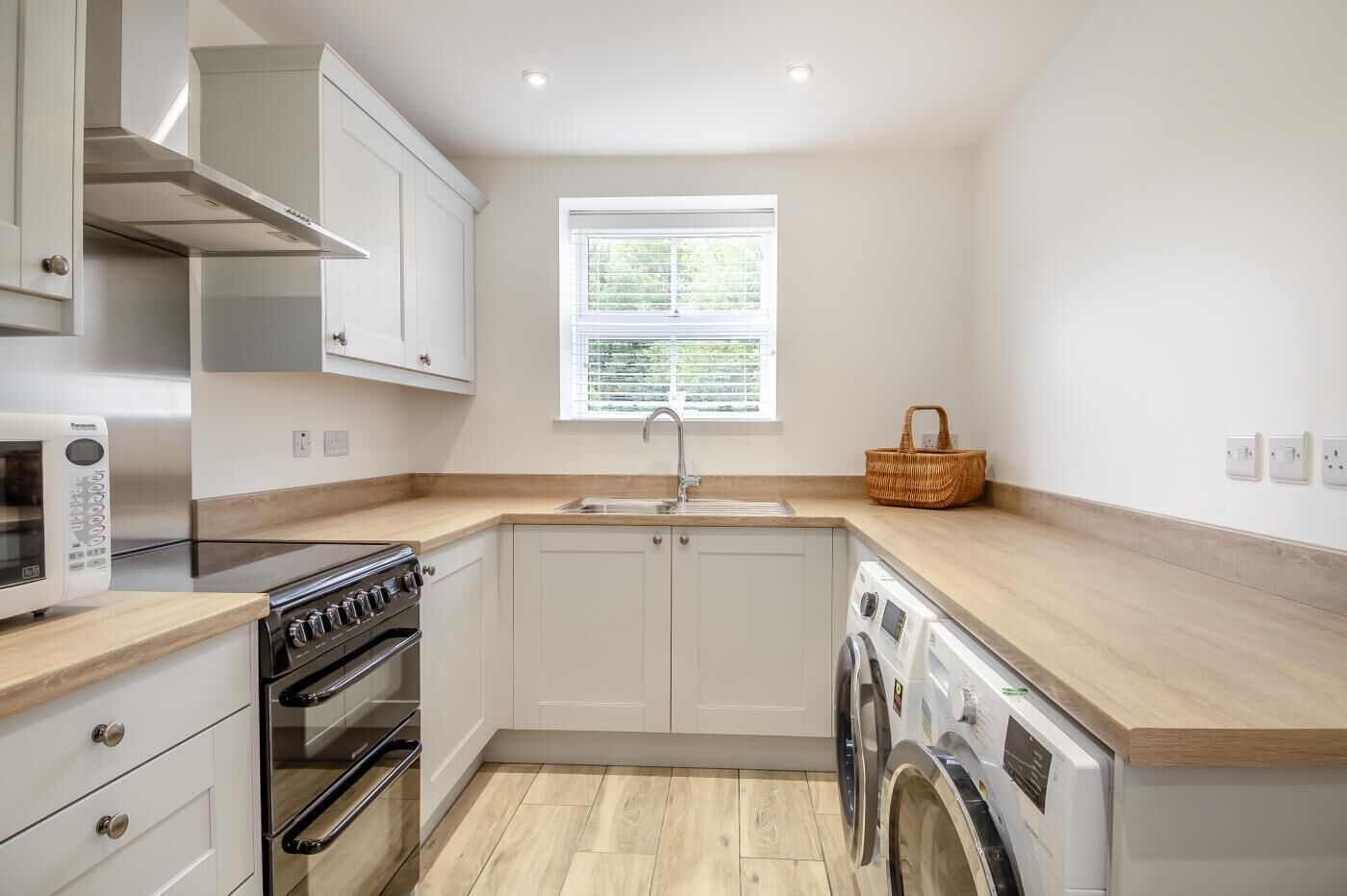 A compact kitchen with white cabinets, a built-in oven, a washing machine, and a window with blinds.