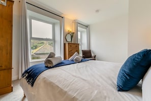 A bedroom with a large bed, a wooden wardrobe, a dresser, and a window with a view of a house. - 2 Bed in Mousehole (oc-d29111) (Mousehole)