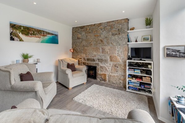 A living room with a stone wall, two armchairs, a fireplace, a TV, and a bookshelf. - 2 Bed in Mousehole (oc-d29111) (Mousehole)
