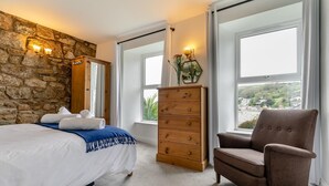 A bedroom with a stone wall, a bed with white linens, a wooden dresser, a brown armchair, and large windows with a view. - 2 Bed in Mousehole (oc-d29111) (Mousehole)
