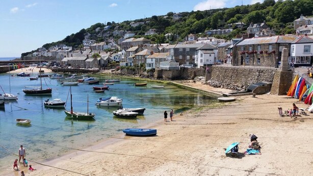 A coastal town with boats docked along the shore, a sandy beach, and houses built on a hillside. - 2 Bed in Mousehole (oc-d29111) (Mousehole)