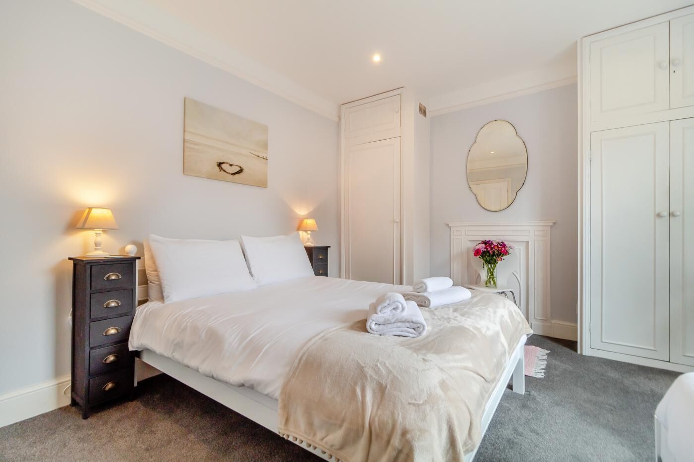 A neatly arranged bedroom with a bed, bedside tables, a dresser, a mirror, and a fireplace.