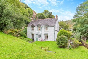 A white house with a gabled roof, surrounded by lush greenery and a hill in the background. - 2 Bed in Boscastle (oc-f29161) (Boscastle)