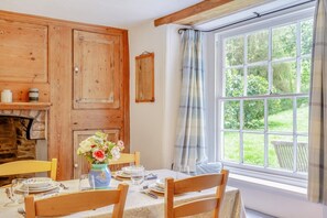 A dining area with a table set for four, wooden cabinets, a fireplace, and a large window with a view of greenery. - 2 Bed in Boscastle (oc-f29161) (Boscastle)