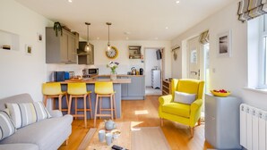 A modern kitchen with a central island, bar stools, a yellow armchair, and a sofa in a bright living area with wooden flooring. - 1 Bed in Minster (oc-l29119) (Boscastle)