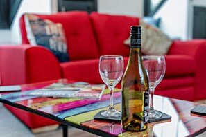 Un sofá rojo, una mesita de vidrio con una botella de vino y dos copas, y revistas coloridas.