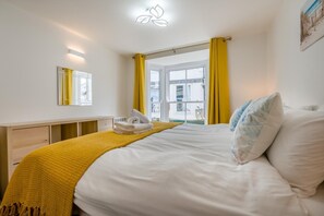 A hotel room with a bed, a bedside table, a mirror, and a window with yellow curtains. - 2 Bed in St. Ives (oc-e28156) (St Ives)