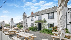 An outdoor seating area with wooden tables and umbrellas, a white building with a black awning, and a garden with flowers and plants. - 4 Bed in St. Merryn (oc-t29153) (Saint Merryn)